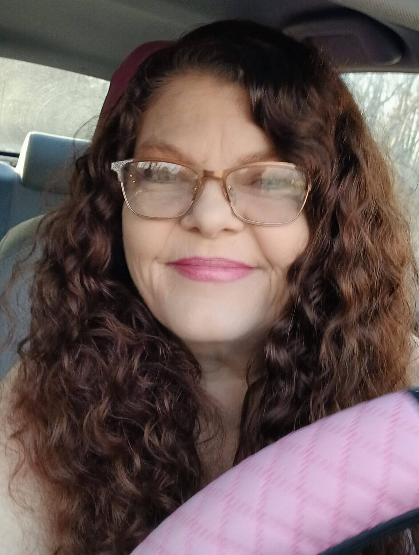 Woman with curly hair and glasses smiling while seated in a vehicle, holding a steering wheel.